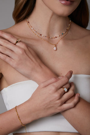 Close-up of a woman wearing gold jewelry including layered necklaces, rings, and bracelet on a neutral background.