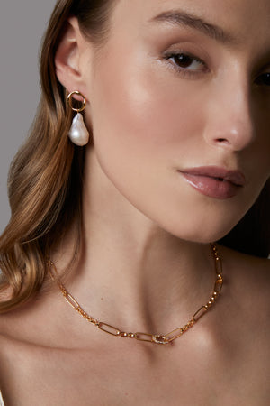 Close-up of a woman wearing a gold necklace and pearl earring against a neutral background