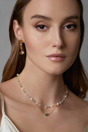 Woman wearing two green gemstone necklaces and leaf earrings against a neutral background