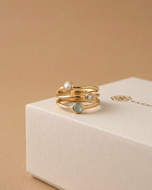 Three gold rings with gemstones on a beige box with 'Karma' branding.