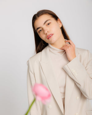A woman wearing a light beige double-breasted blazer over a matching beige mock-neck top, wearing large skinny hammered gold hoop earrings with a hand-hammered texture