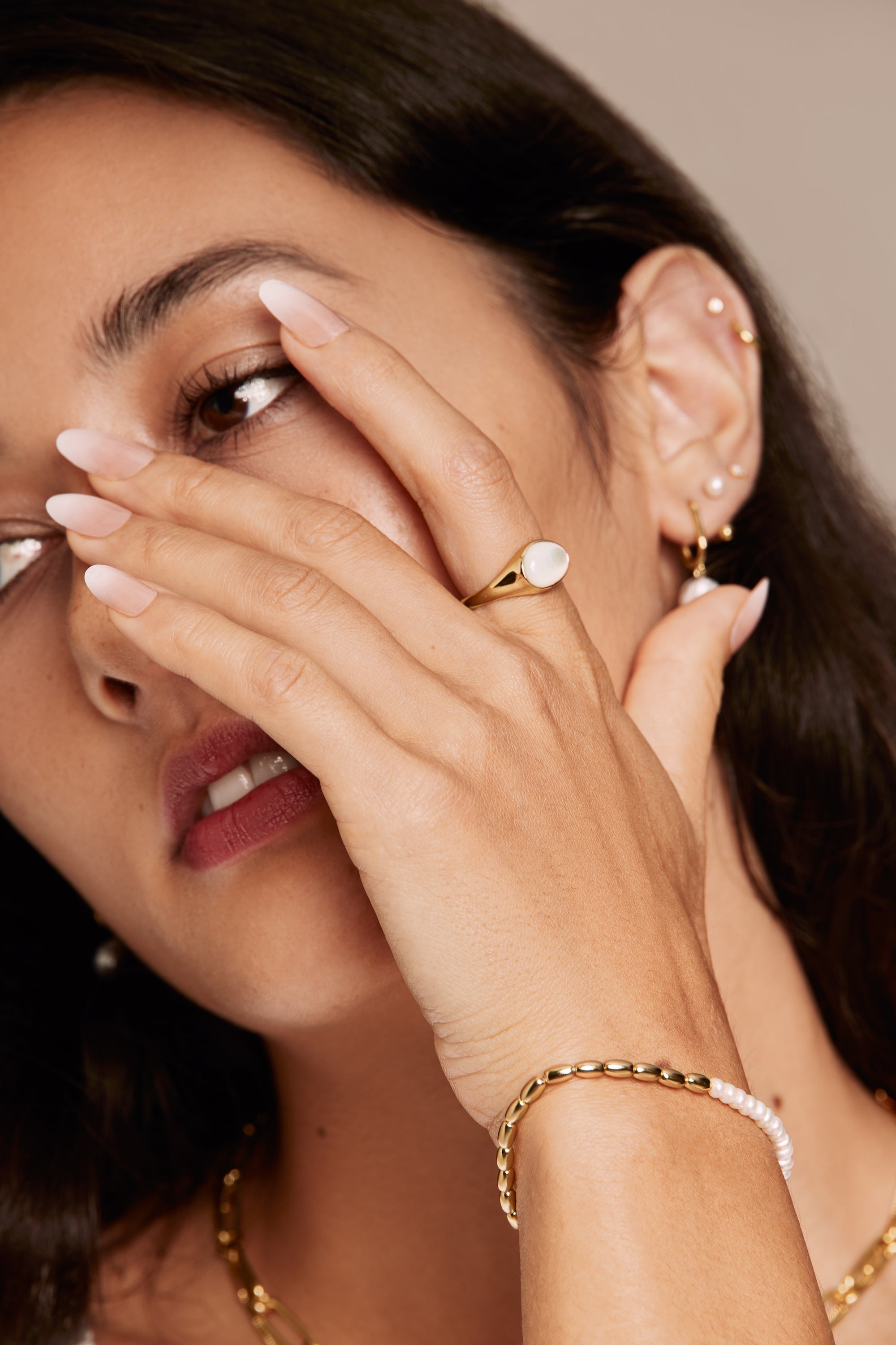 Close-up of a woman wearing gold jewelry including layered necklaces, ring, and pearl bracelet on a neutral background.