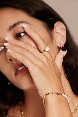 Close-up of a woman wearing gold jewelry including layered necklaces, ring, and pearl bracelet on a neutral background.
