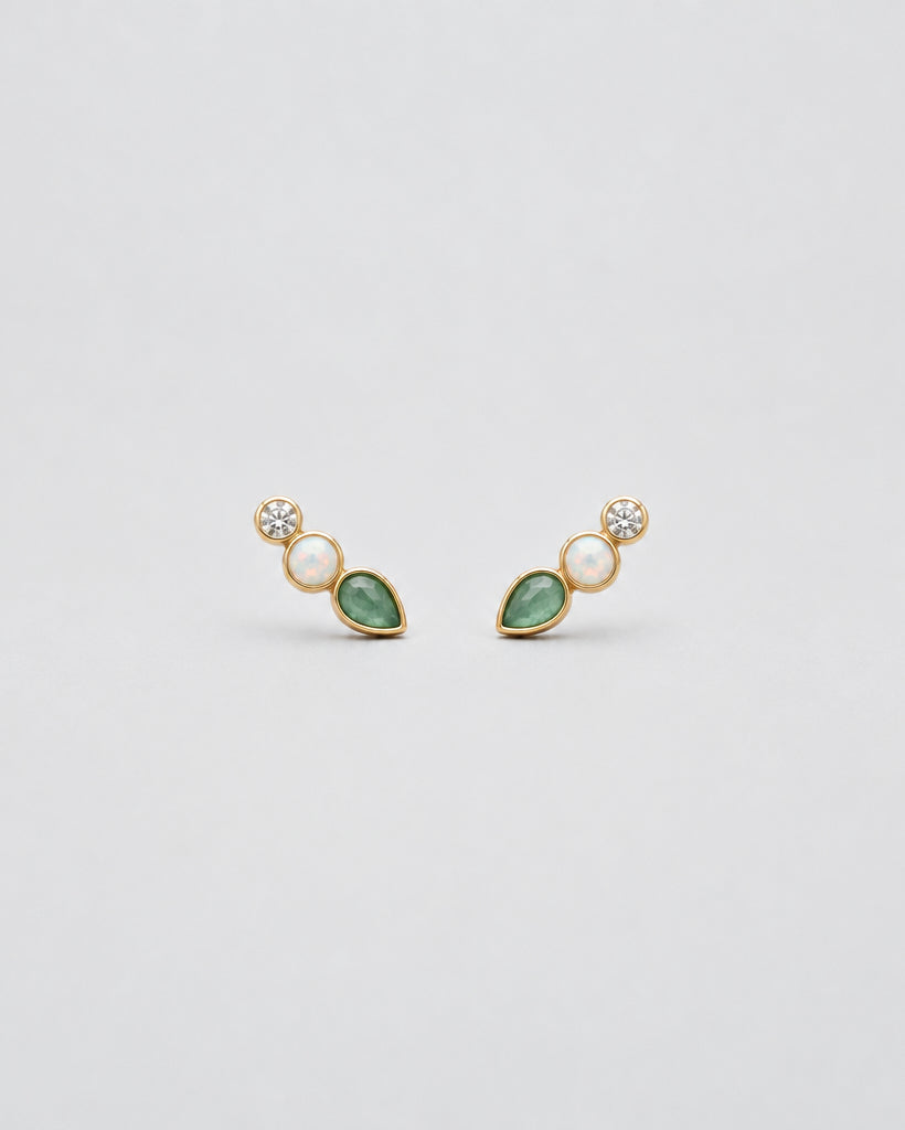 A pair of small gold-toned stud earrings on a textured off-white background. Each earring features a round clear stone, a small oval pale pink stone, and a teardrop-shaped green stone.