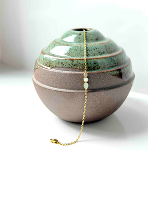 A gold chain anklet with three small light-colored beads and a tiny clear stone, draped over a ceramic vase with a green glazed top and a speckled brown base.