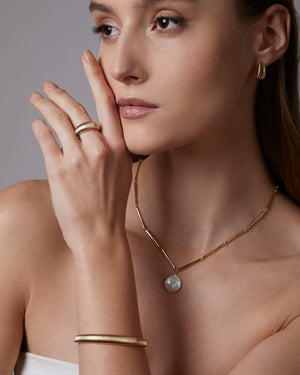 Woman wearing gold jewelry including a necklace, ring, and dome cuff bracelet against a neutral background