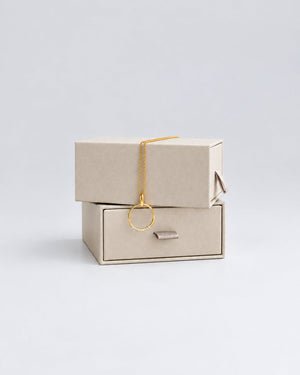 A delicate gold necklace with a single circular ring pendant, draped over stacked beige jewelry boxes on a light neutral background.