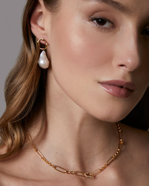 Close-up of a woman wearing a gold necklace and pearl earring against a neutral background