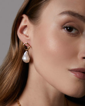 Close-up of a woman wearing gold hoop earrings with a baroque pearl drop and a gold chain necklace.