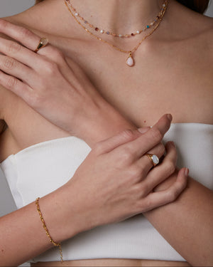 Close-up of a woman wearing gold jewelry including a necklace, ring, and oval bracelet on a neutral background.
