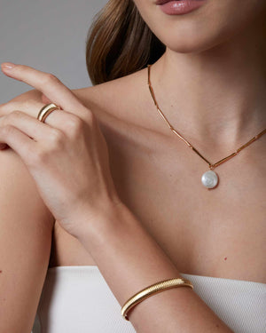 Woman wearing gold jewelry with a pearl coin necklace, and gold brass dome bangle bracelet and a ring on a neutral background