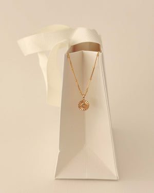 Gold necklace with a sun pendant on a white paper bag  against a beige background