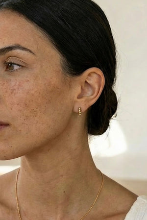 Woman wearing a gold necklace and beaded huggie hoop earrings with a neutral background