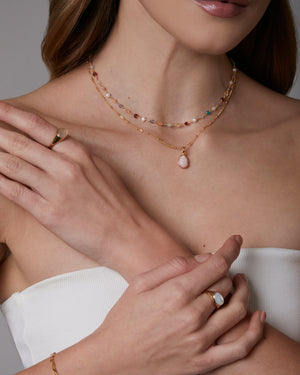 Close-up of a woman wearing gold jewelry including layered necklaces, rings, and bracelet on a neutral background.