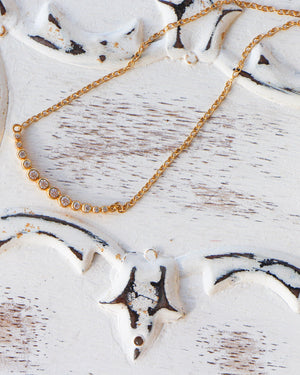 A gold-plated sterling silver necklace with a curved bar charm adorned with cubic zirconia stones, displayed on a white textured surface.